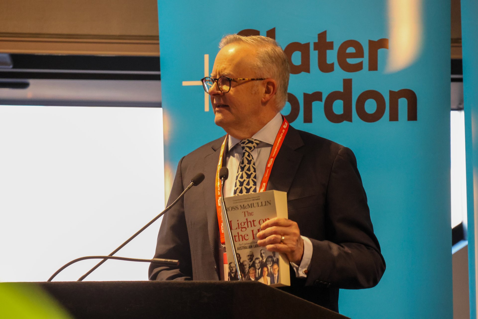 Anthony Albanese w okularach i garniturze przemawia przy mównicy, trzyma w ręku książkę