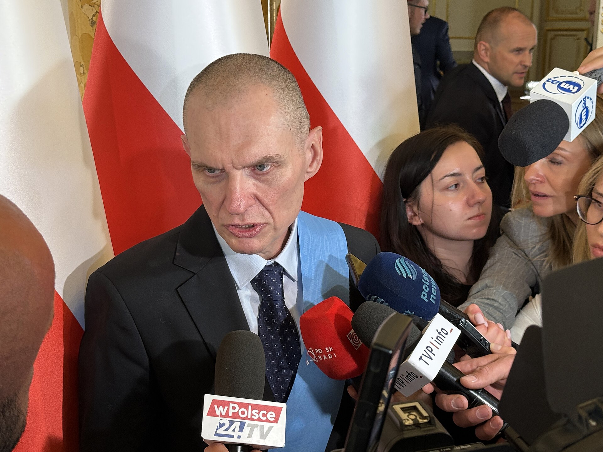 Mężczyzna z niebieską wstęgą orderu na piersi odpowiada na pytania dziennikarzy, przed nim mikrofony stacji telewizyjnych, za nim flagi Polski