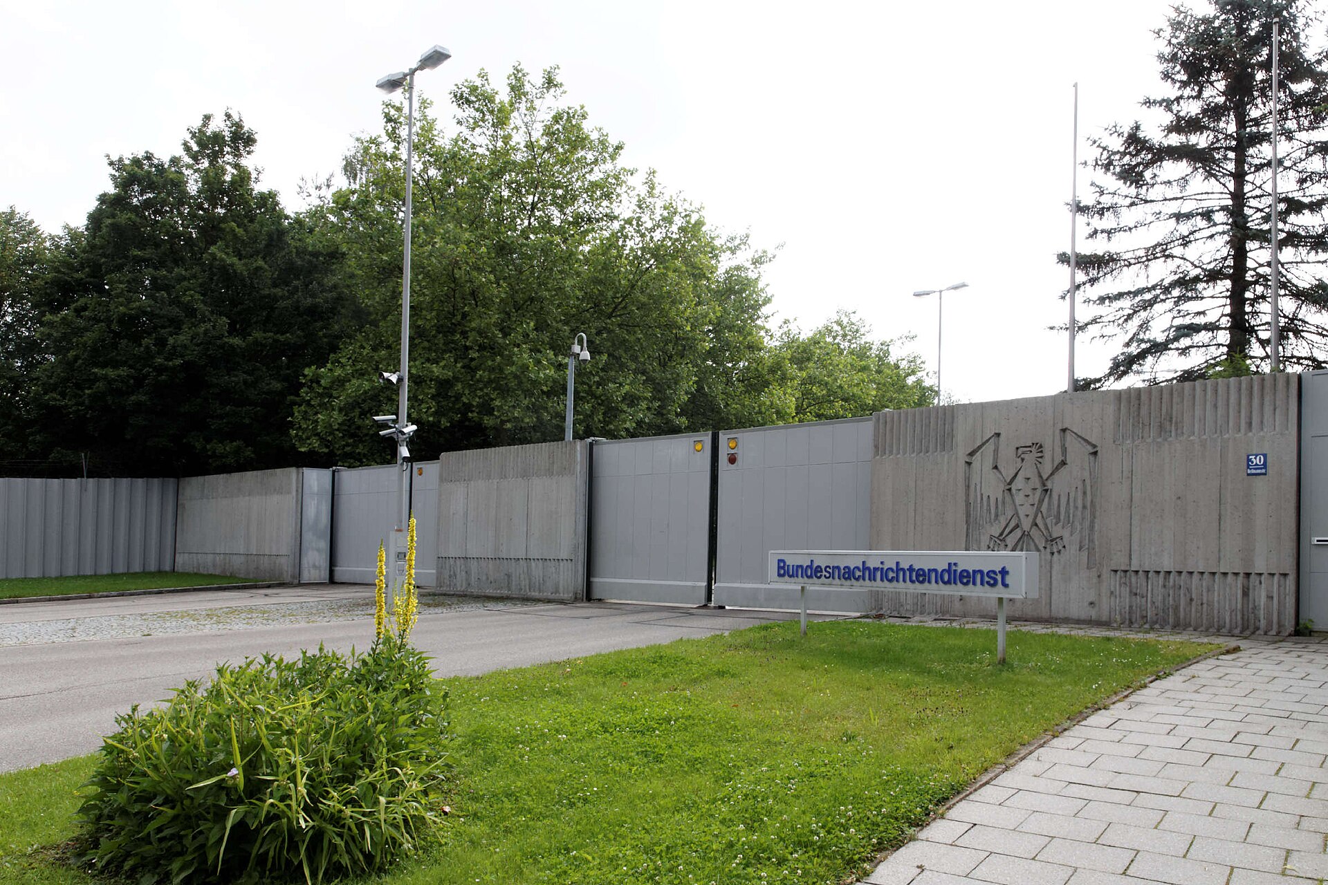 Szara metalowa brama i mur z wizerunkiem orła, przed nią tablica z napisem Bundesnachrichtendienst, obok trawnik i lampy