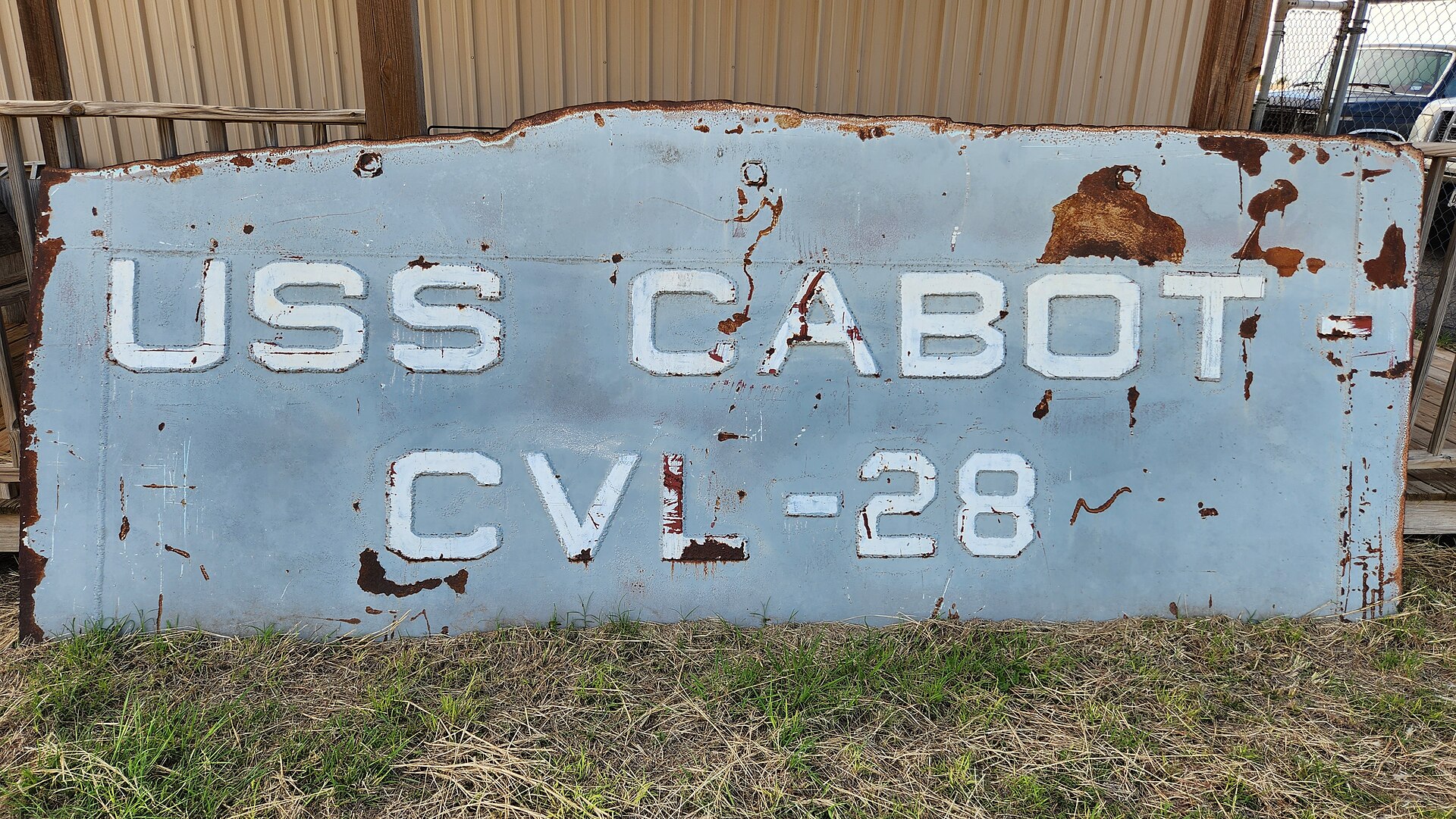 Zardzewiała niebieska blacha z białymi literami USS Cabot CVL-28 stoi oparta o ścianę na trawie