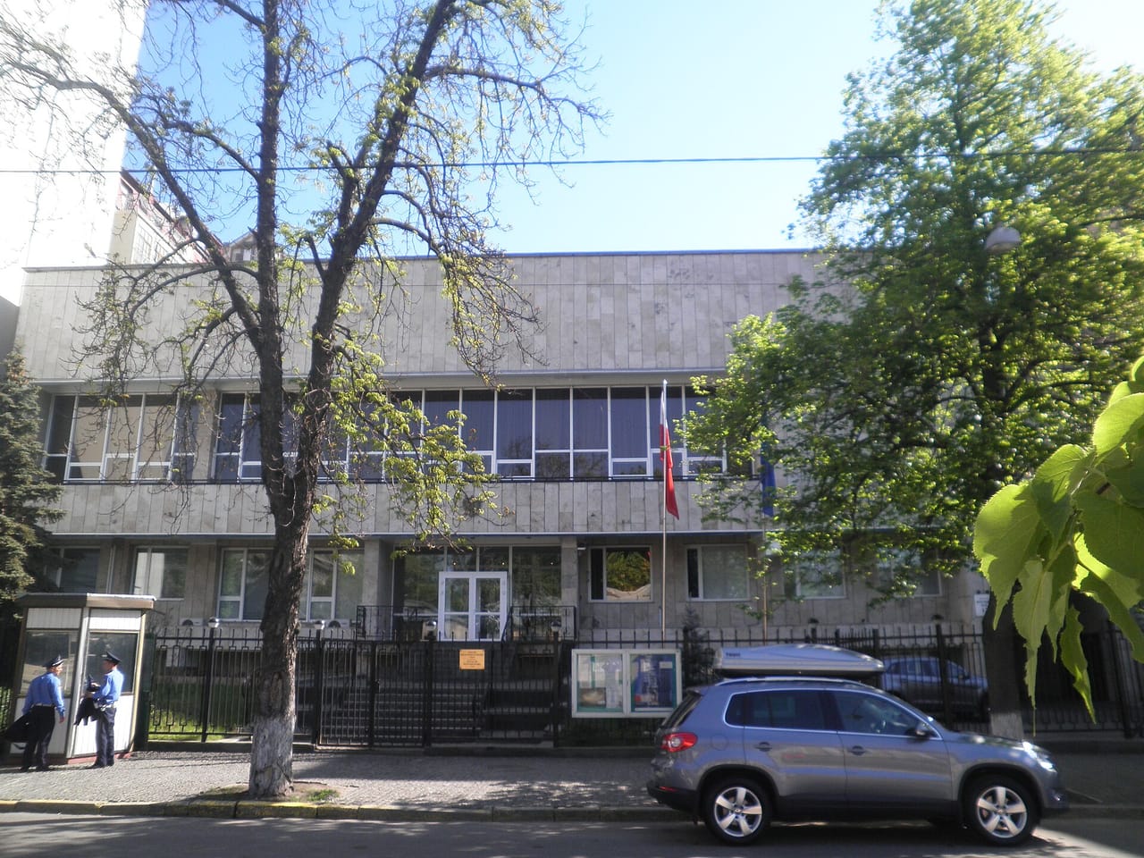 Dwupiętrowy budynek obłożony jasnym kamieniem, przed nim ogrodzenie, budka strażnicza i polska flaga na maszcie