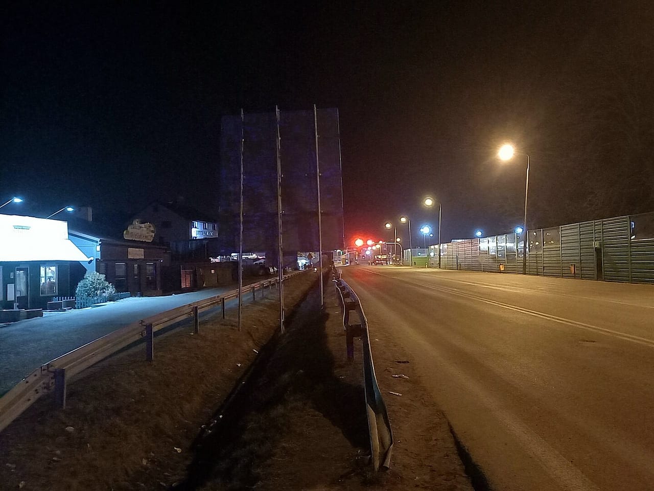 Oświetlona latarniami droga do przejścia granicznego nocą, po prawej zielone ekrany, w oddali czerwone światła