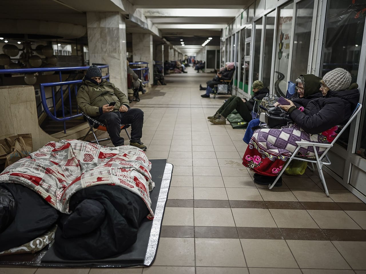 Ludzie siedzą na krzesełkach i leżą pod kocami w podziemnym korytarzu stacji metra