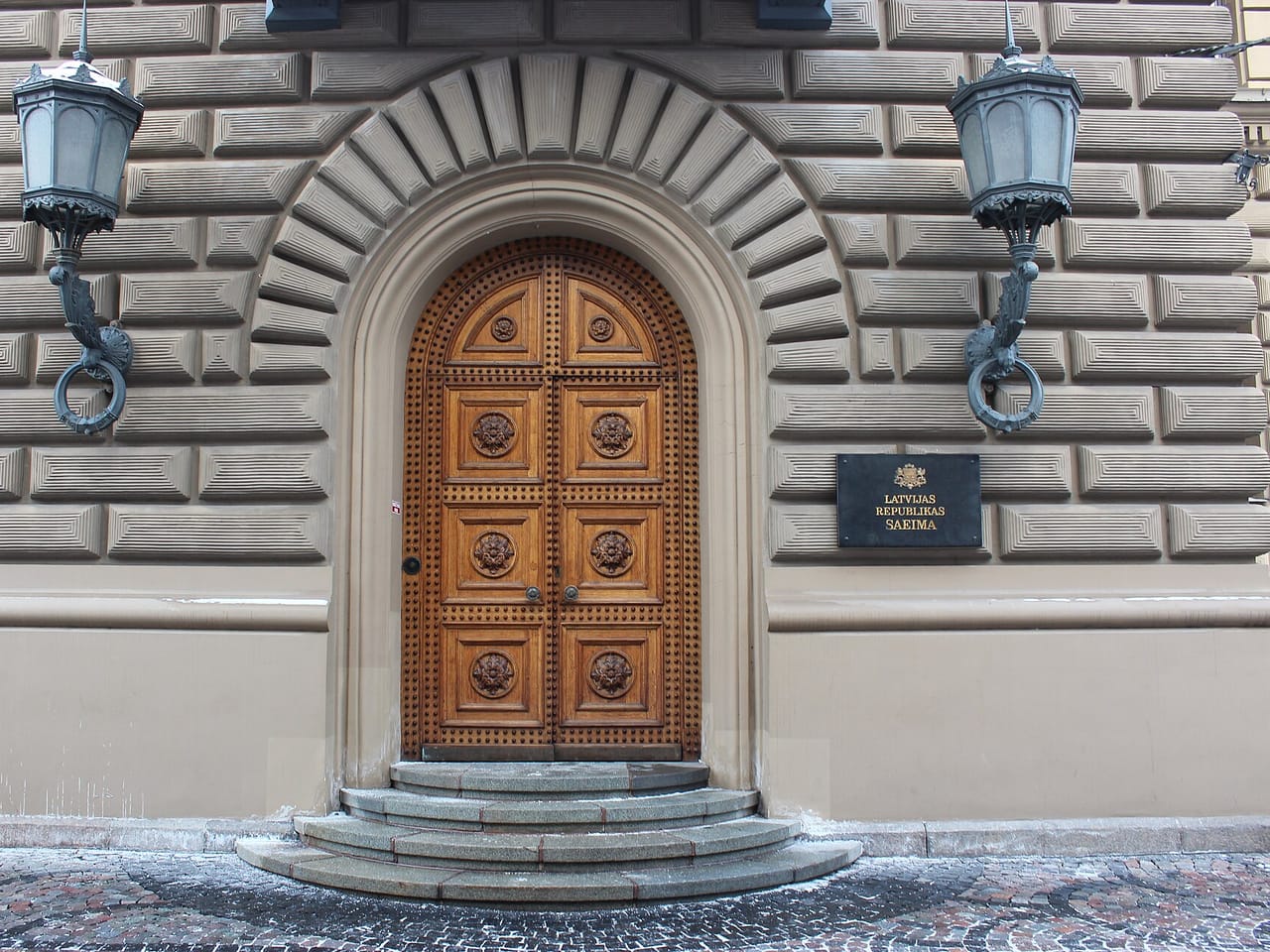 Drewniane łukowe drzwi w kamiennej fasadzie, po bokach dwie latarnie, obok tablica z napisem Latvijas Republikas Saeima
