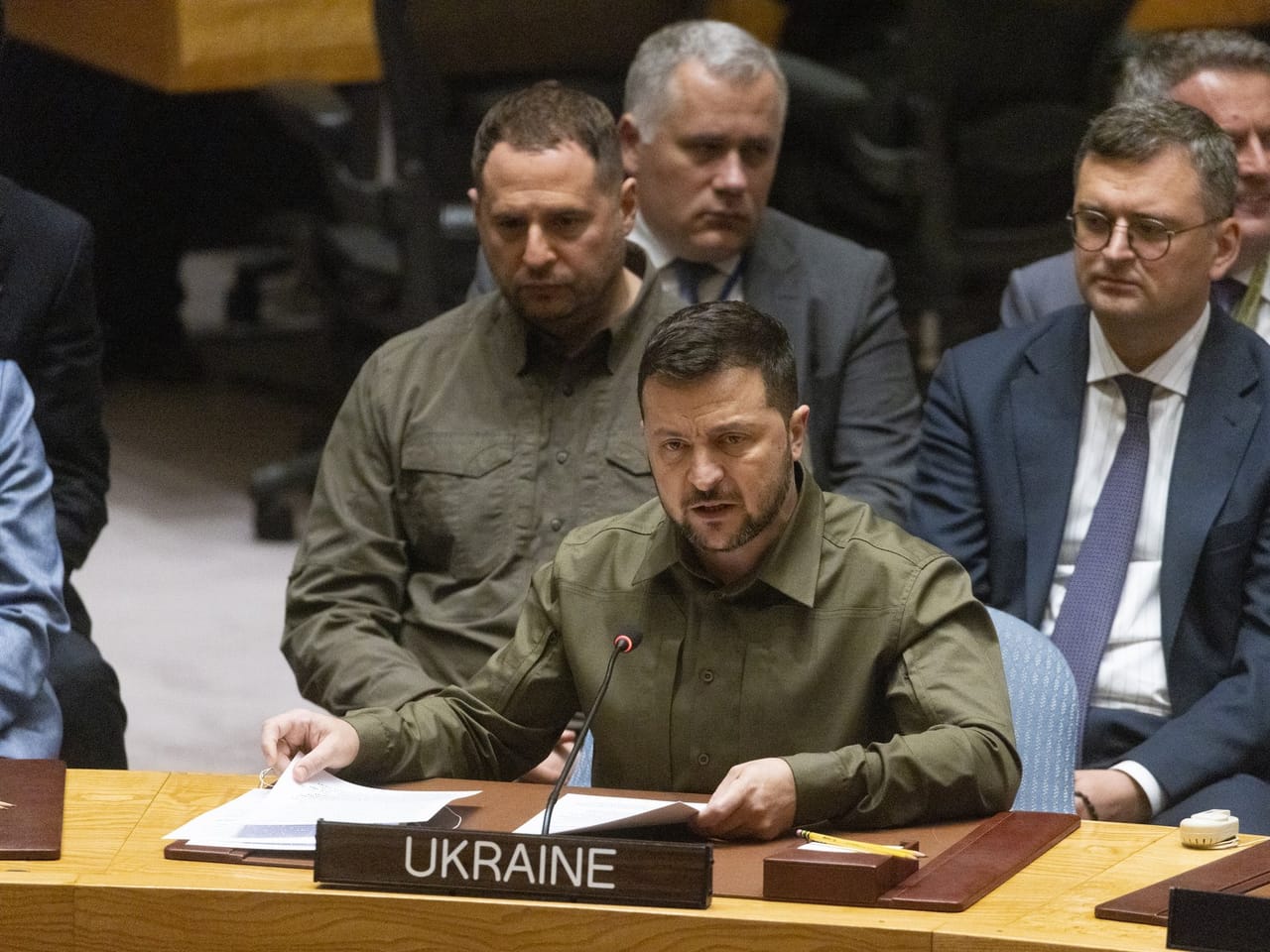 Wołodymyr Zełenski w oliwkowej koszuli przemawia przy stole z tabliczką Ukraine, za nim członkowie delegacji