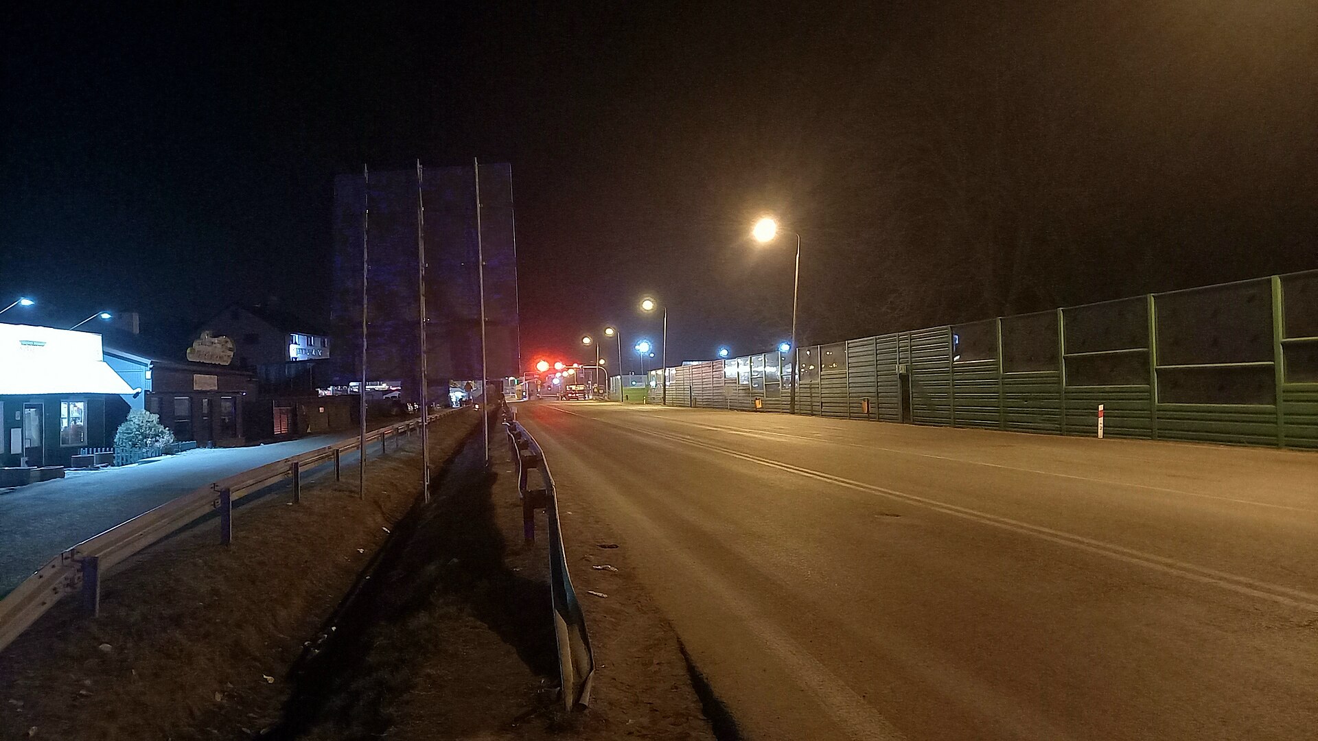 Oświetlona latarniami droga do przejścia granicznego nocą, po prawej zielone ekrany, w oddali czerwone światła