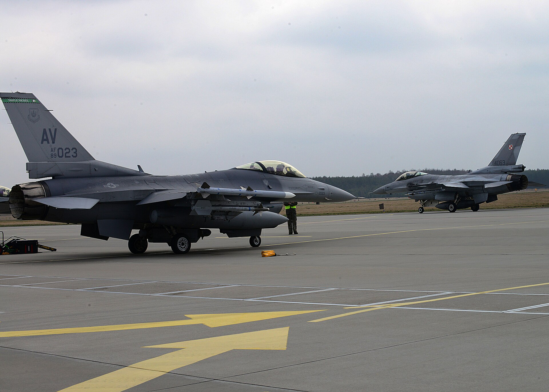 Dwa myśliwce F-16 na płycie lotniska, z przodu amerykański, w tle polski z szachownicą na stateczniku
