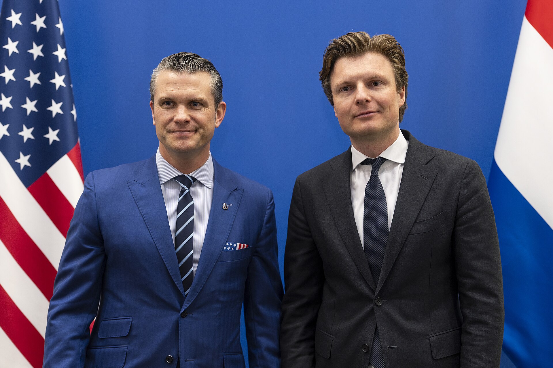 Pete Hegseth i holenderski minister obrony Ruben Brekelmans pozują przed niebieskim tłem z flagami USA i Holandii