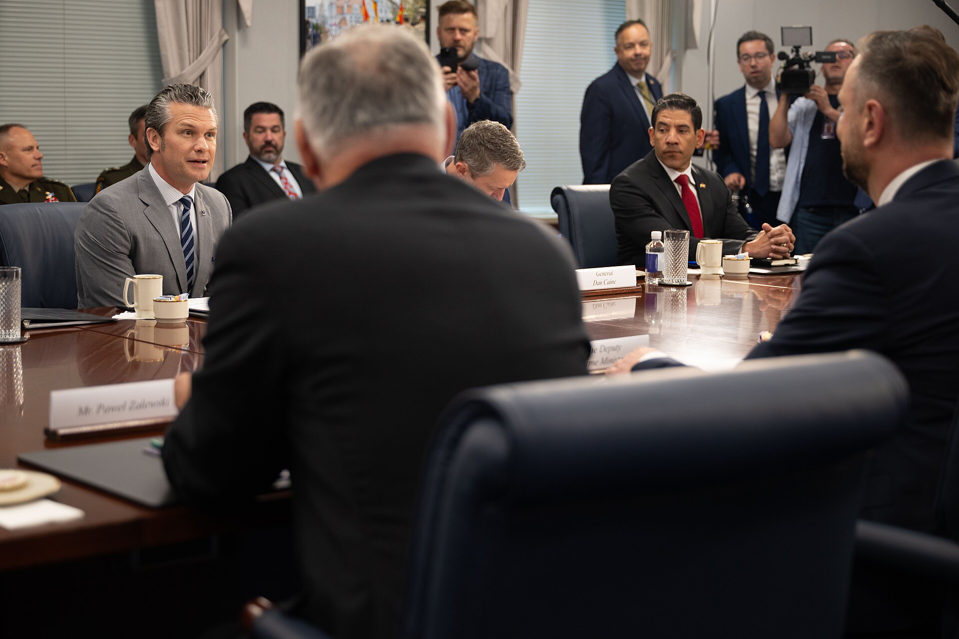 Mężczyźni w garniturach siedzą przy długim stole konferencyjnym, po lewej mówi Pete Hegseth, w tle fotoreporterzy