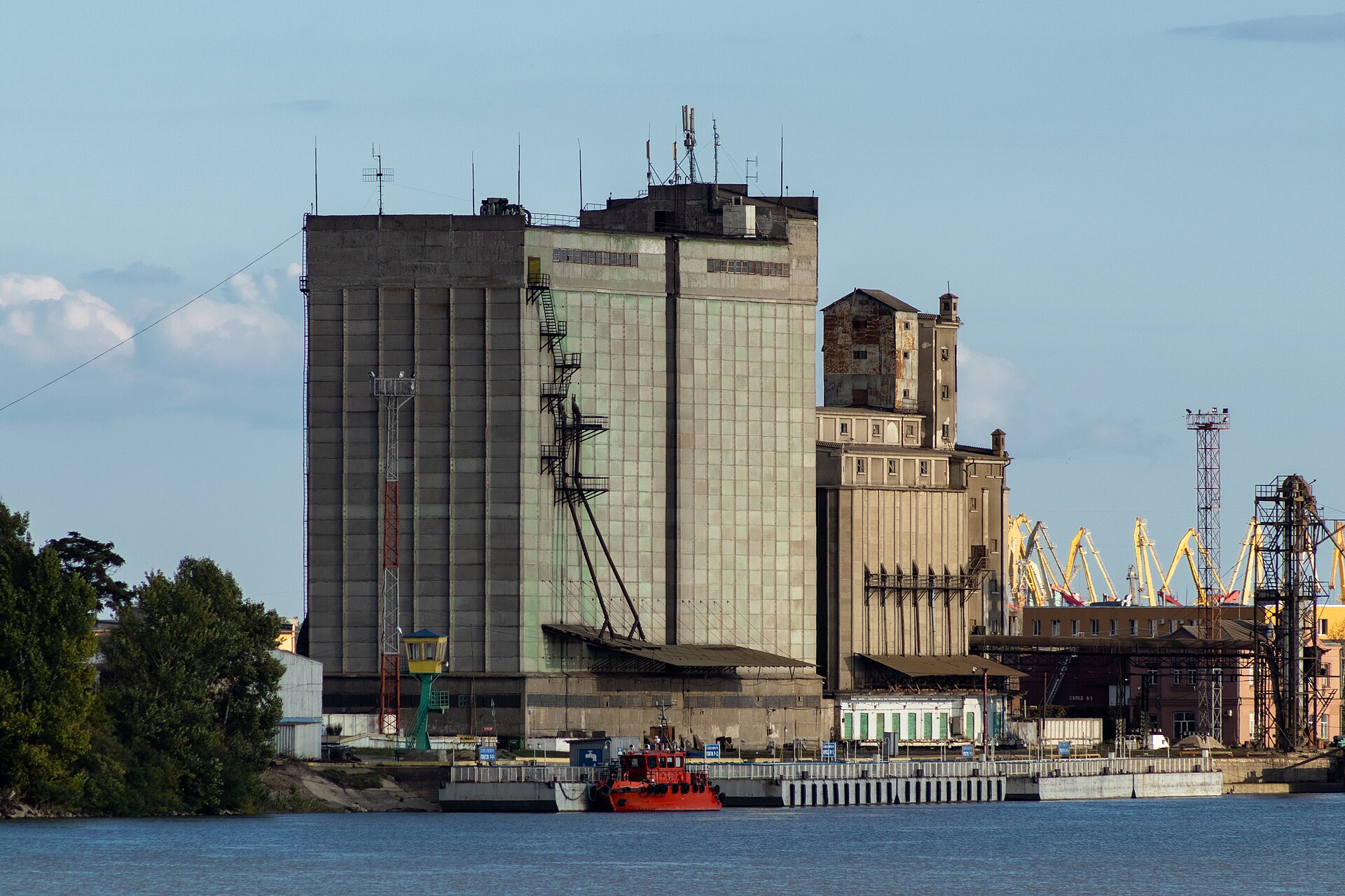 Wielki betonowy elewator zbożowy nad rzeką, przy nabrzeżu czerwony holownik, w tle żółte dźwigi portowe