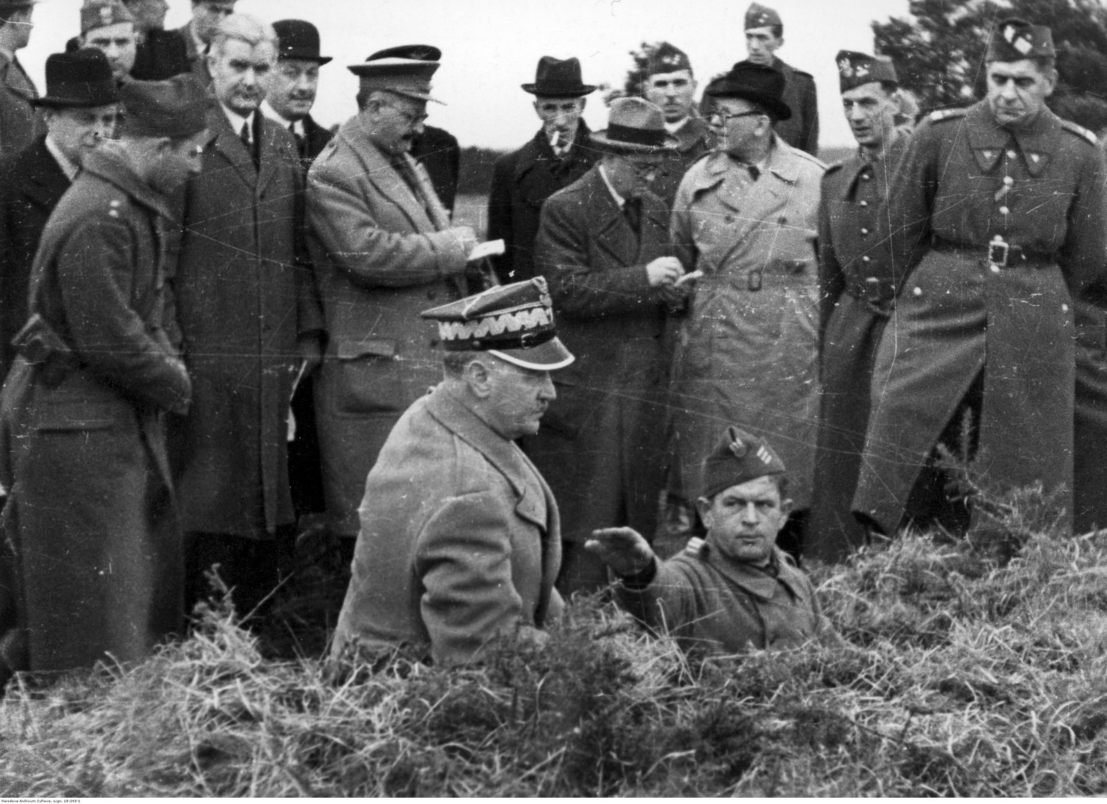 Gen. Władysław Sikorski w rogatywce generalskiej obserwuje ćwiczenia z okopu, za nim grupa oficerów i cywilów