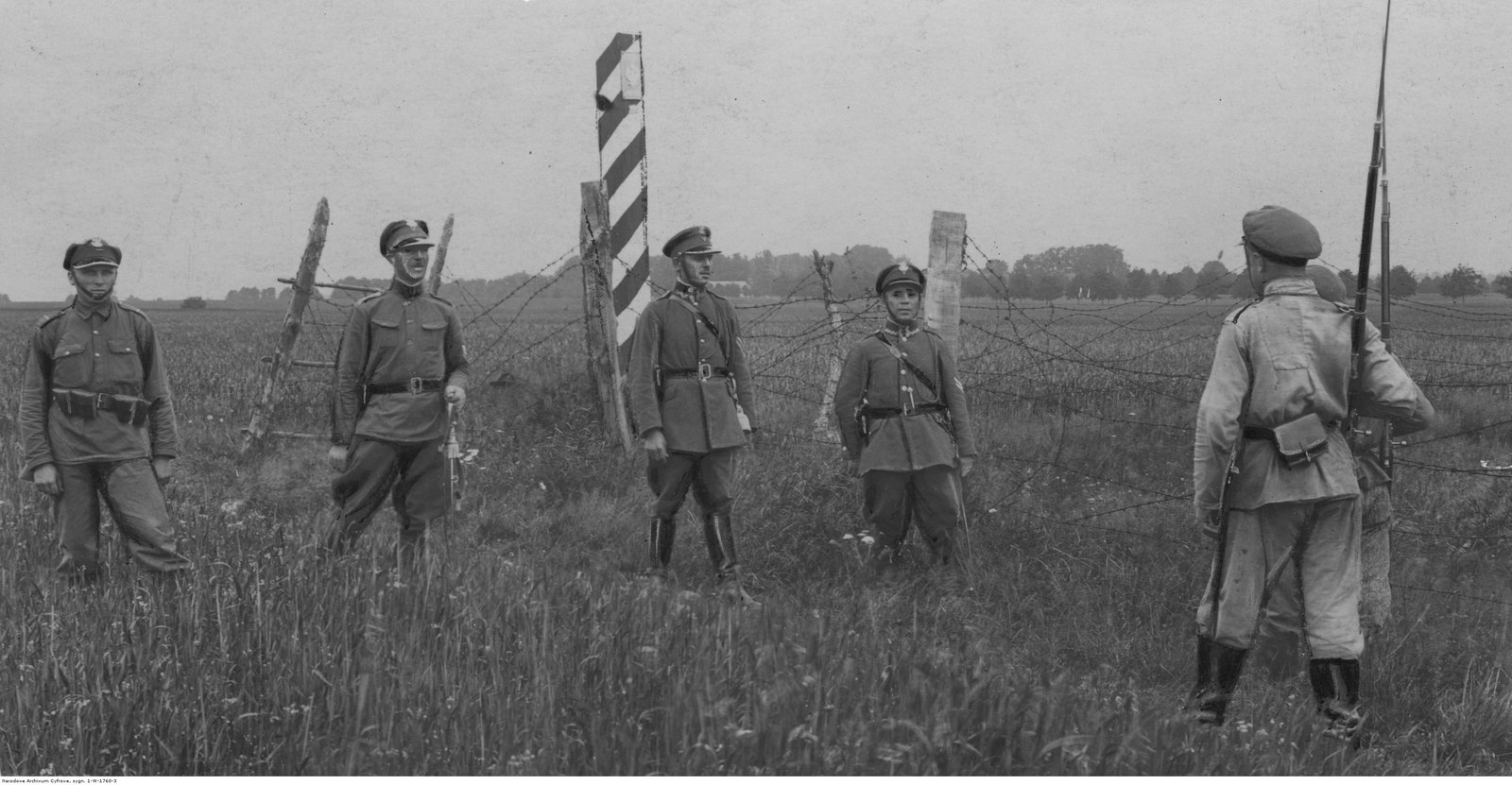 Czterej żołnierze Korpusu Ochrony Pogranicza w rogatywkach stoją przy słupie granicznym i zasiekach naprzeciw dwóch żołnierzy sowieckich z karabinami