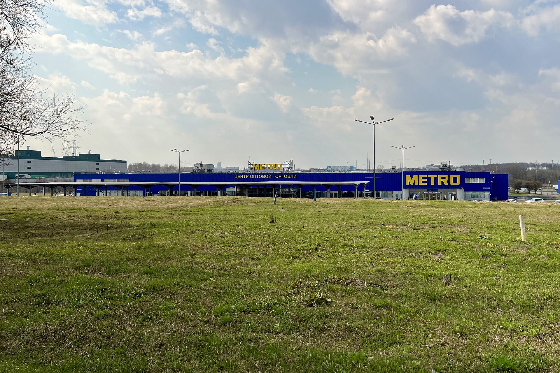Niska niebiesko-żółta hala hurtowni z napisem Metro za trawiastym polem, nad nią chmury