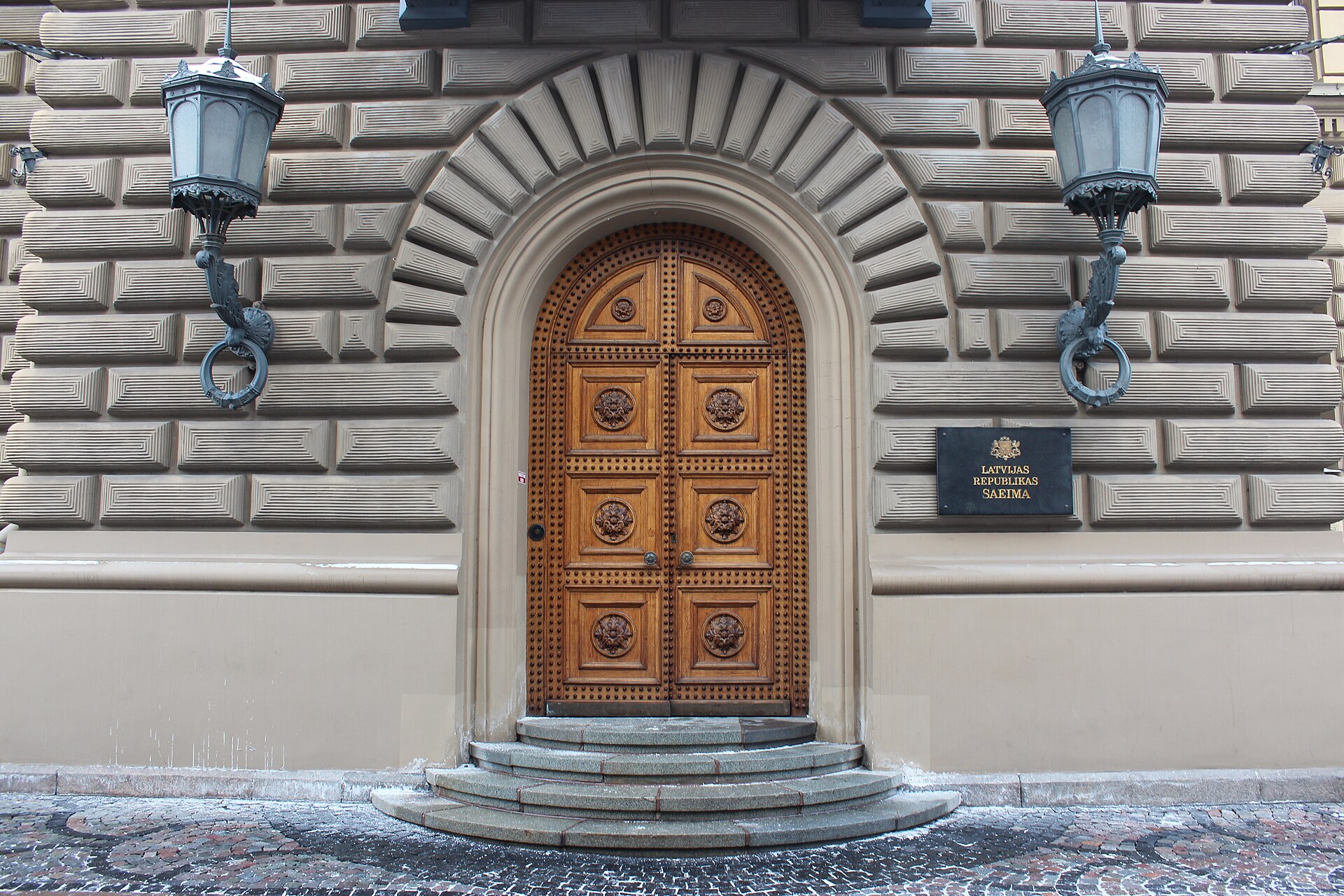 Drewniane łukowe drzwi w kamiennej fasadzie, po bokach dwie latarnie, obok tablica z napisem Latvijas Republikas Saeima
