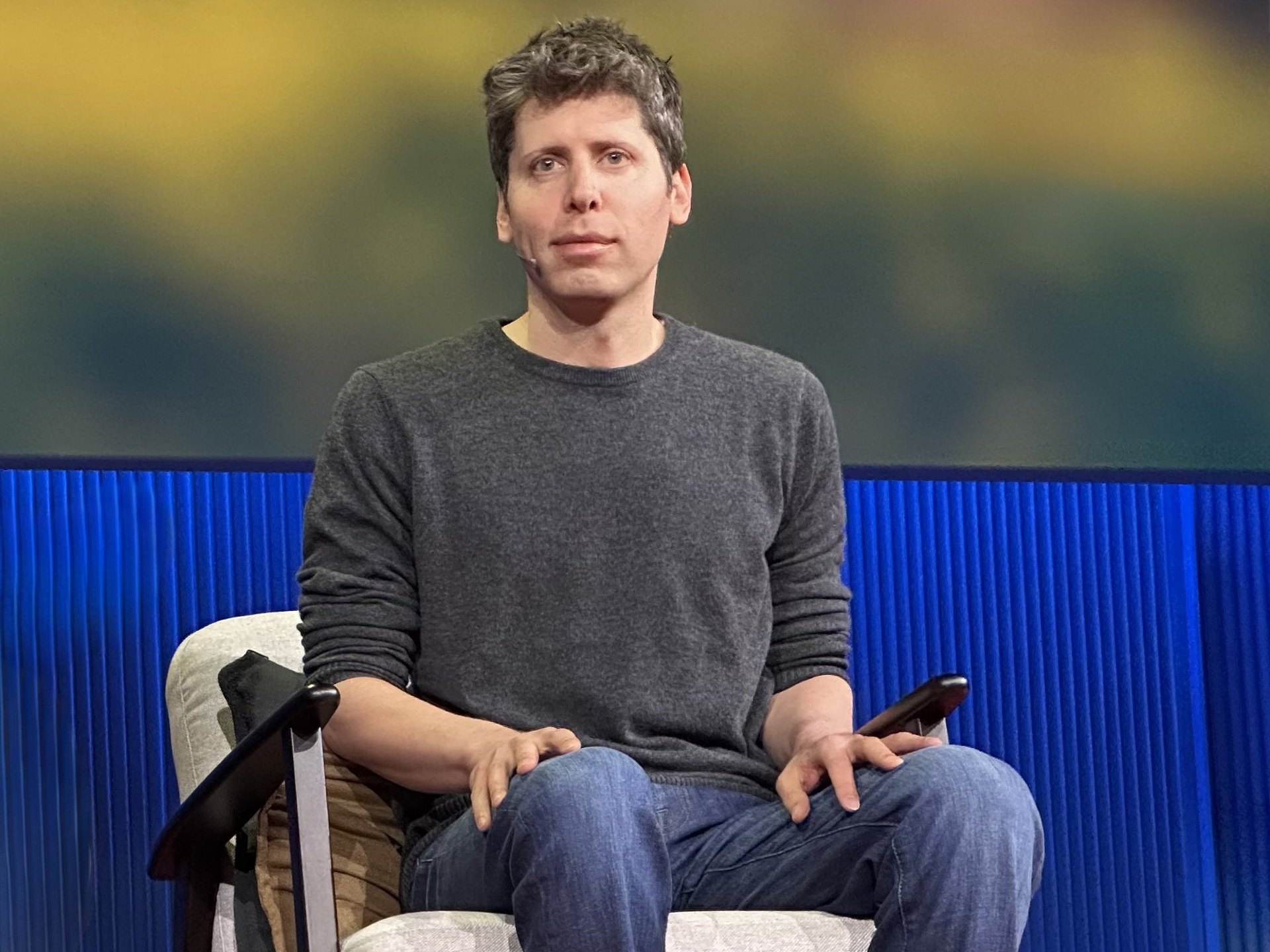 Sam Altman w szarym swetrze siedzi w fotelu na scenie z niebieskim tłem