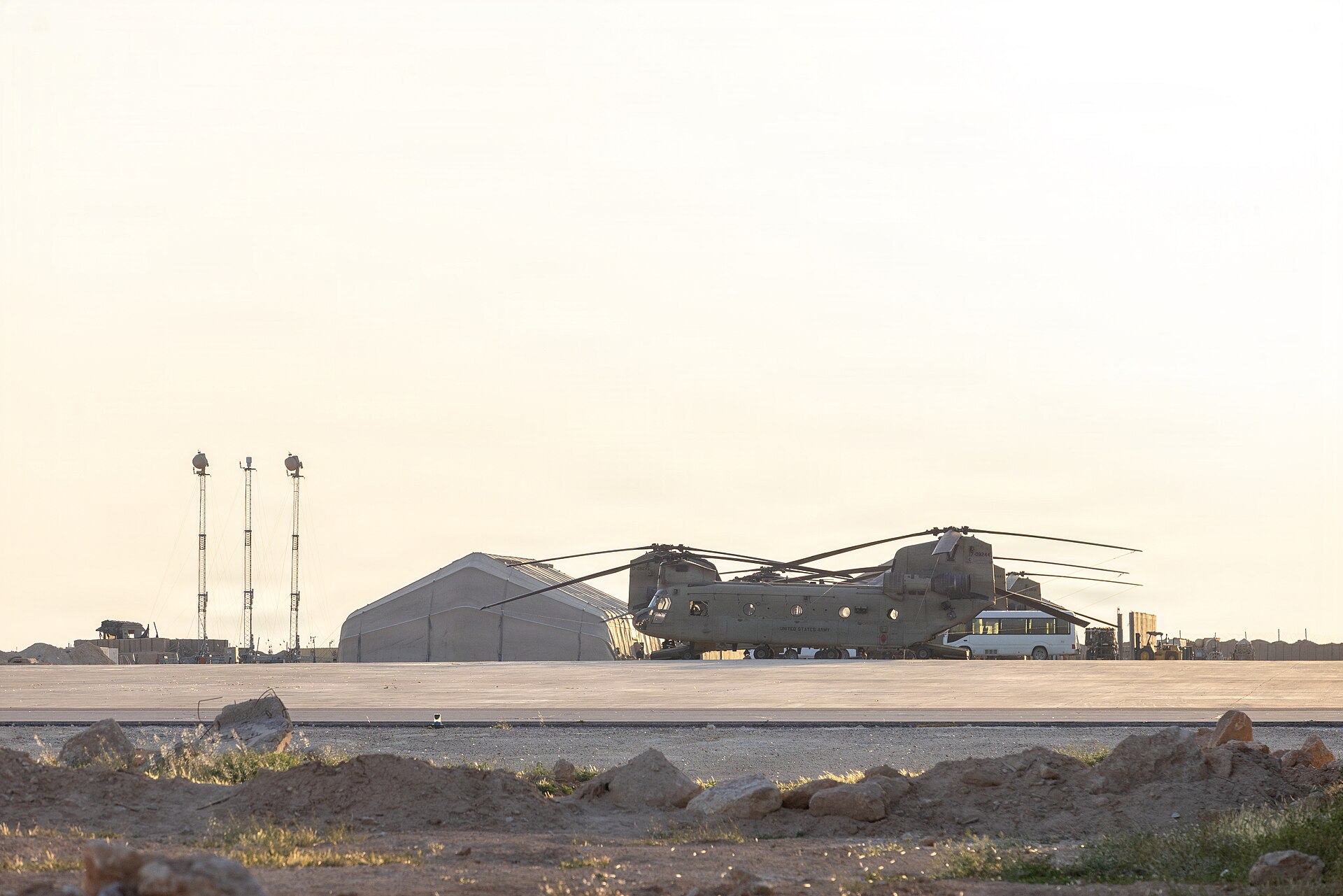 Dwa śmigłowce transportowe Chinook stoją na płycie lotniska przed namiotowym hangarem, w tle wieże oświetleniowe i blade niebo