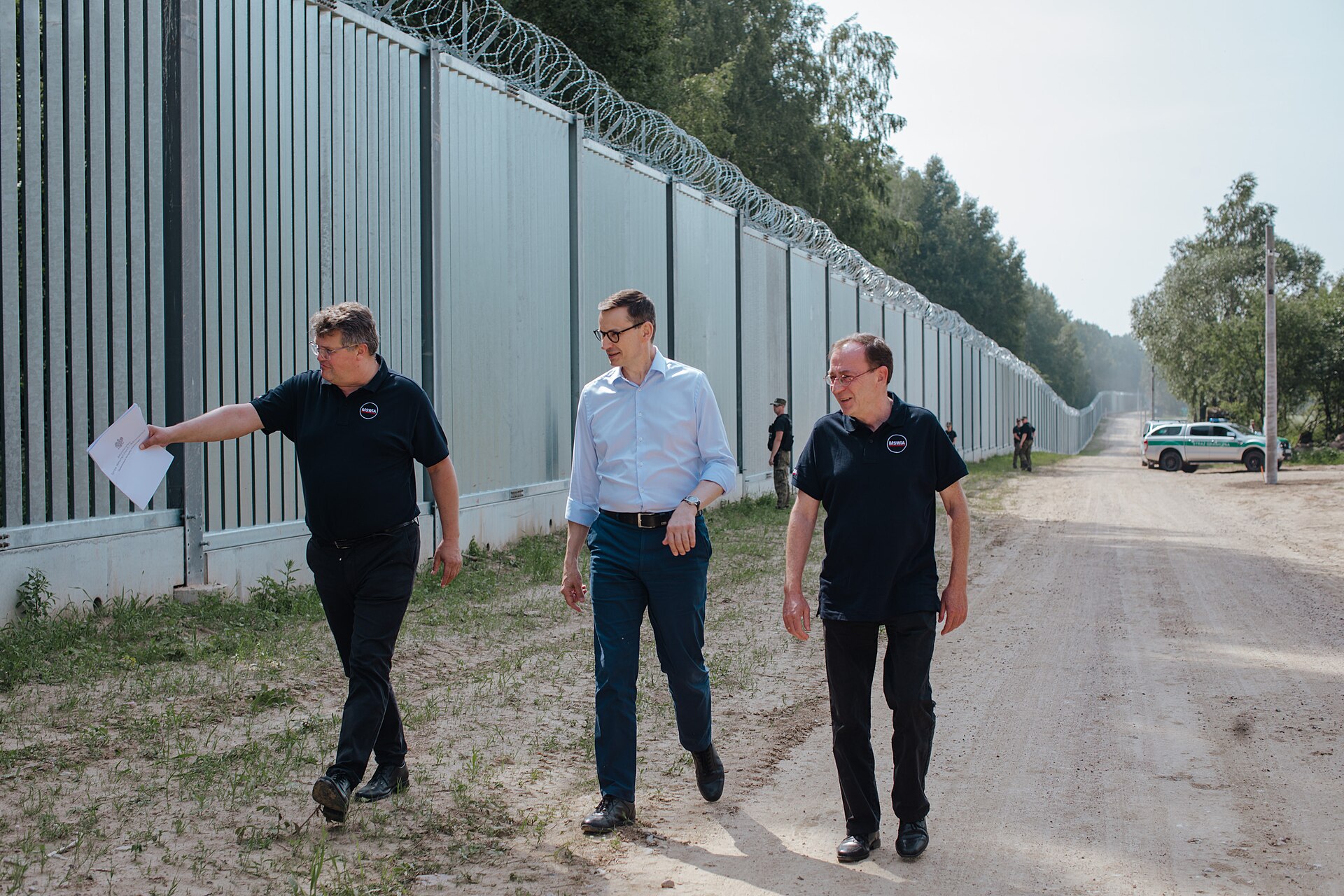 Trzech mężczyzn idzie drogą gruntową wzdłuż wysokiej stalowej zapory zwieńczonej drutem kolczastym