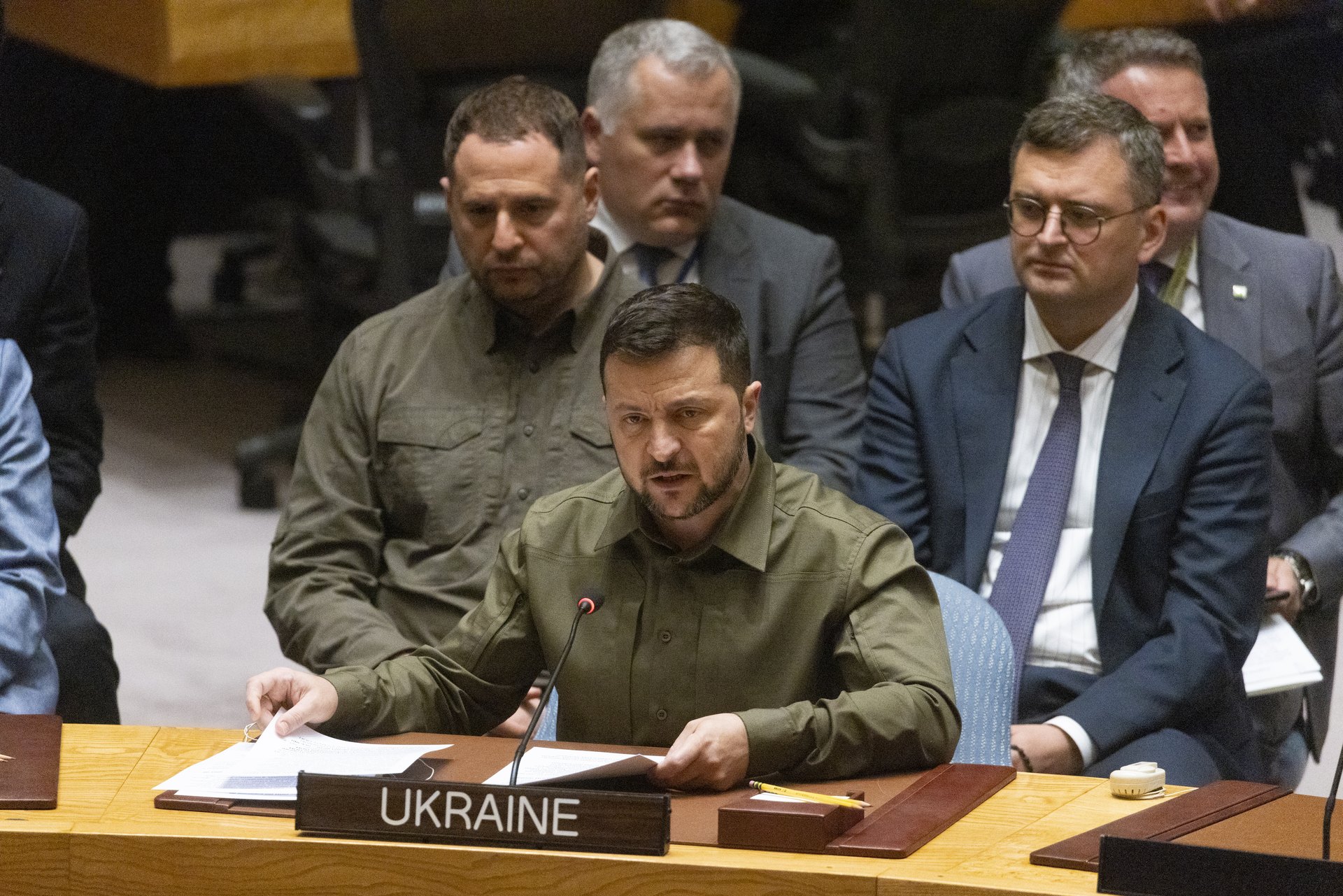 Wołodymyr Zełenski w oliwkowej koszuli przemawia przy stole z tabliczką Ukraine, za nim członkowie delegacji